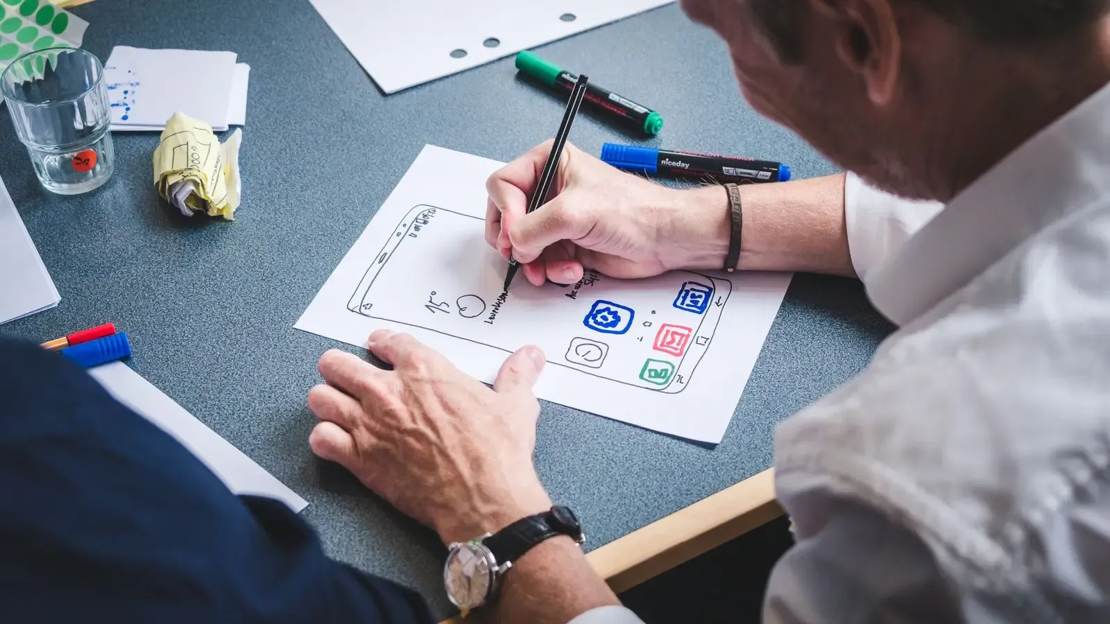 Person zeichnet mit Marker einen App-Prototyp auf Papier, daneben liegen Skizzen und Stifte